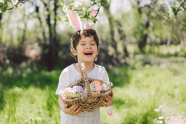 Забавный мальчик с корзиной яиц и заячьими ушами на пасхальных яйцах в солнечном весеннем саду — стоковое фото