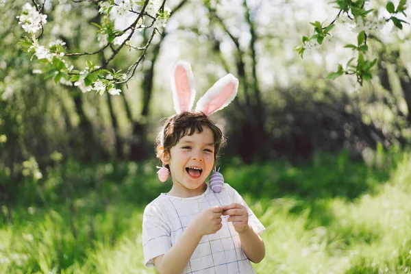 Смішний хлопчик з яєчним кошиком і вухами кролика на полюванні на великодні яйця в сонячному весняному саду — стокове фото
