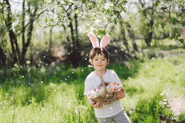 Vicces fiú tojáskosár és nyuszifül húsvéti tojás vadászat a napos tavaszi kertben — Stock Fotó