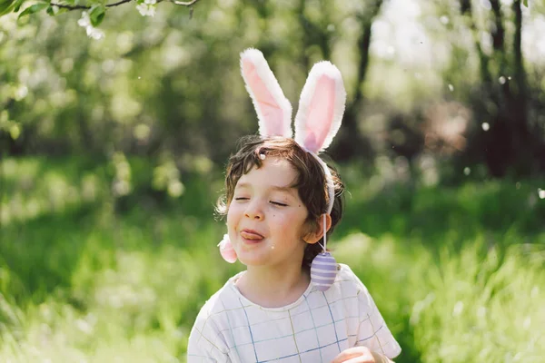Смішний хлопчик з яєчним кошиком і вухами кролика на полюванні на великодні яйця в сонячному весняному саду — стокове фото