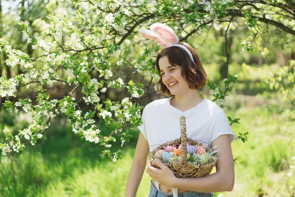 Смешная девочка-подросток с корзиной яиц и заячьими ушами на пасхальных яйцах в солнечном весеннем саду — стоковое фото