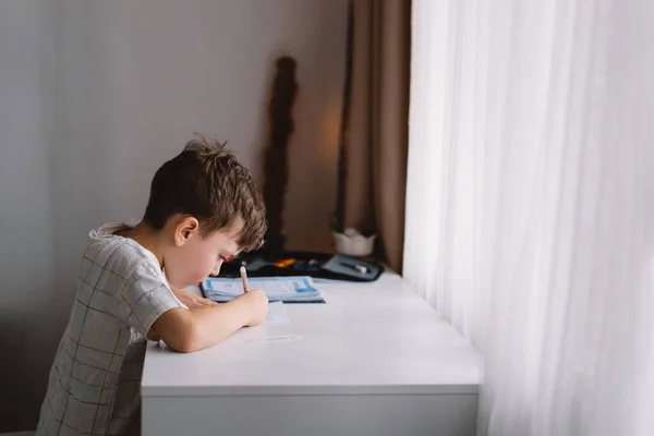 Aranyos kisfiú, aki otthon tanul és házi feladatot csinál. Gondolkozó gyermek ül az asztalnál. — Stock Fotó