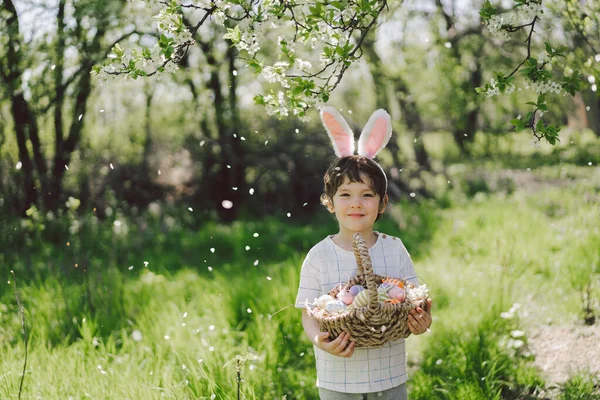 Vicces fiú tojáskosár és nyuszifül húsvéti tojás vadászat a napos tavaszi kertben — Stock Fotó