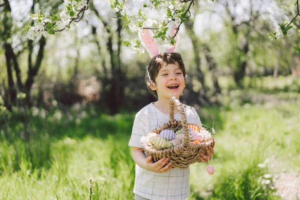 Забавный мальчик с корзиной яиц и заячьими ушами на пасхальных яйцах в солнечном весеннем саду — стоковое фото