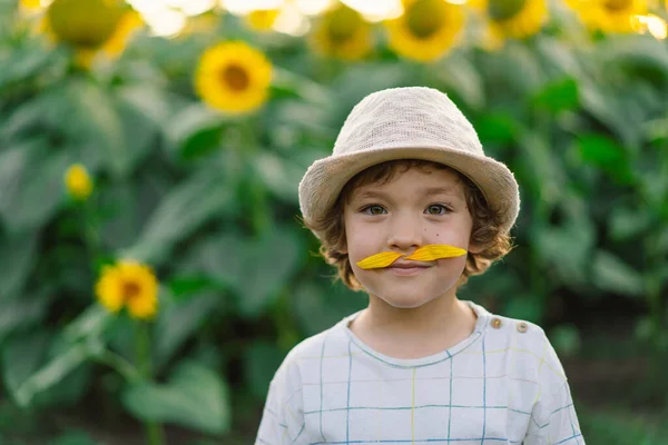 Lycklig liten pojke gå i fält av solrosor och göra en mustasch av solrosor. — Stockfoto