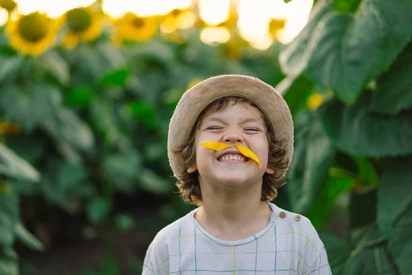 Lycklig liten pojke gå i fält av solrosor och göra en mustasch av solrosor. — Stockfoto