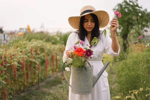 Beautiful brunette girl in a white dress and hat with watering can watering flowers — Stock Photo, Image
