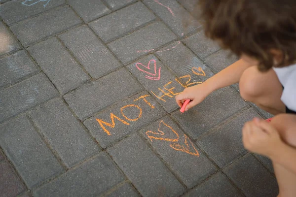 Rapaz bonito desenha a palavra mãe com giz. O menino ama sua mãe. — Fotografia de Stock