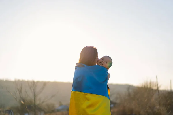 Жінка обіймає свого сина, загорнутого в жовтий і синій прапор України на відкритому повітрі.. — стокове фото
