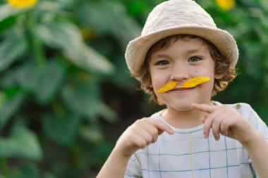 Happy little boy walking in field of sunflowers and making a mustache from sunflower petals. clipart