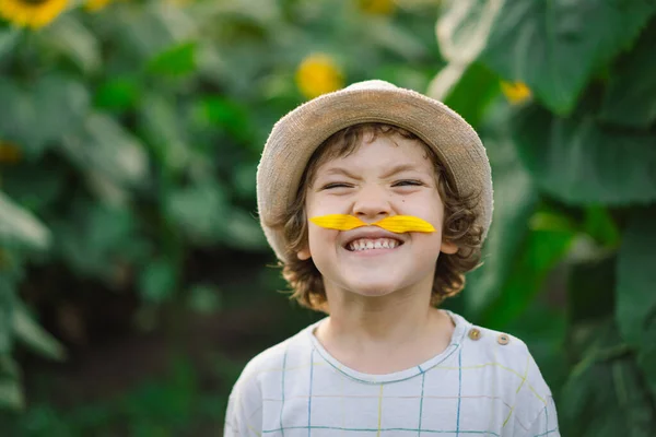 Gelukkig jongetje wandelen in het veld van zonnebloemen en het maken van een snor van zonnebloemblaadjes. — Stockfoto
