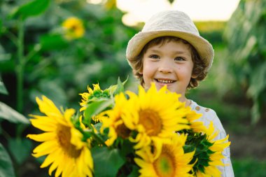 Happy boy walking in field of sunflowers. Child playing with big flower and having fun. clipart