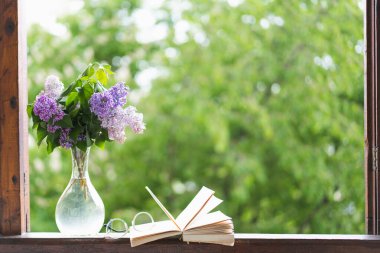 Ahşap bir pencerede kitap, gözlük ve leylak.