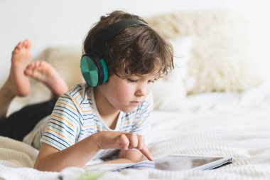Evdeki yatakta uzanıp bilgisayar tabletleriyle oynayan, internetten kitap okuyan ve kablosuz kulaklıkla müzik dinleyen tatlı çocuk.