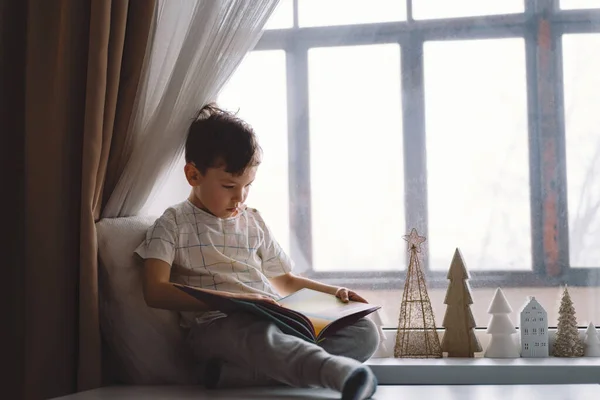 Cute boy is reading a book sitting on the windowsill near the window. Cozy home with decorative Christmas trees. Sweet home. Winter holidays lifestyle.