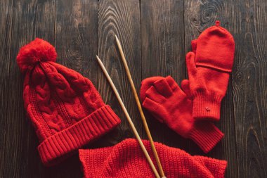 Knitted red hat, scarf, knitted mittens and wooden knitting needles on wooden background. Hobby craft. Concept of knitting, needlework.