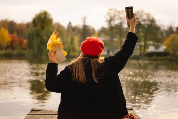 Woman in a red beret reading book on wooden pontoon. Autumn season. Female with trendy clothes relaxing near water reading book and drink coffee. Autumn mood. Autumn concept. Autumn day.