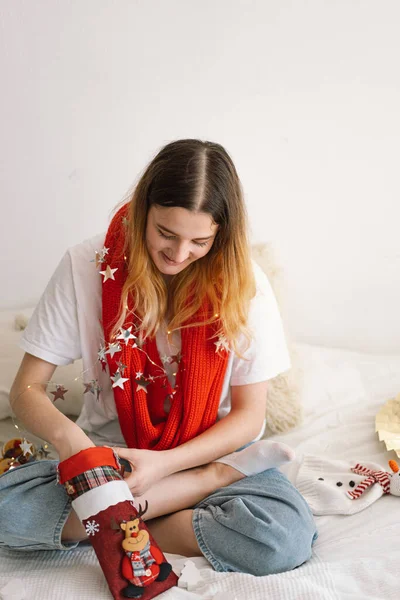 A beautiful girl holds a Christmas stocking in her hands and takes something out of it. Cozy comfortable interior design. Work at home. New year and Christmas concept.