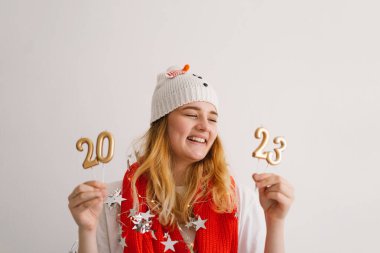 Yeni yıl şapkası ve kırmızı eşarplı komik genç kız 2023 rakamlarını elinde tutuyor. Yeni yıl ve Noel konsepti.
