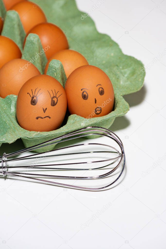 box of chicken eggs with painted faces and a whisk on white background, vertical top view