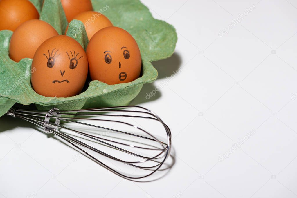 box of chicken eggs with painted faces and a whisk on white background, closeup