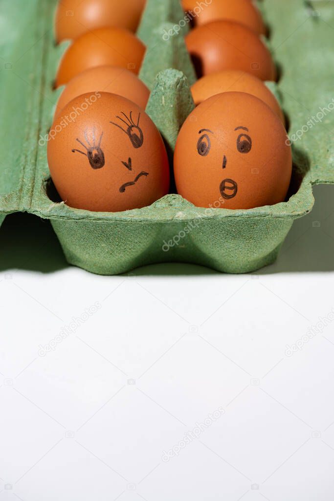 box with chicken eggs with painted faces on white background, vertical top view