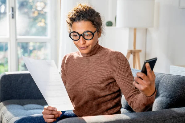 Zwarte Vrouw Lezen Document Het Houden Van Smartphone Thuis — Stockfoto