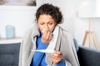 Black woman suffering cold and flu at home checking thermometer