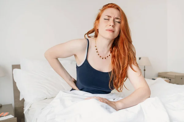 Young woman sitting in bed and suffering from back pain