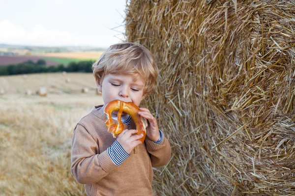Cute toddler eating German pretzel on goden hay field — Stock Photo, Image