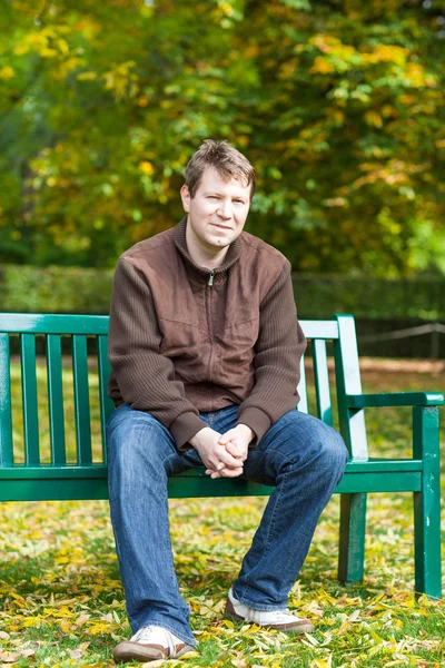 Молодой человек в осеннем парке сидит на скамейке — стоковое фото