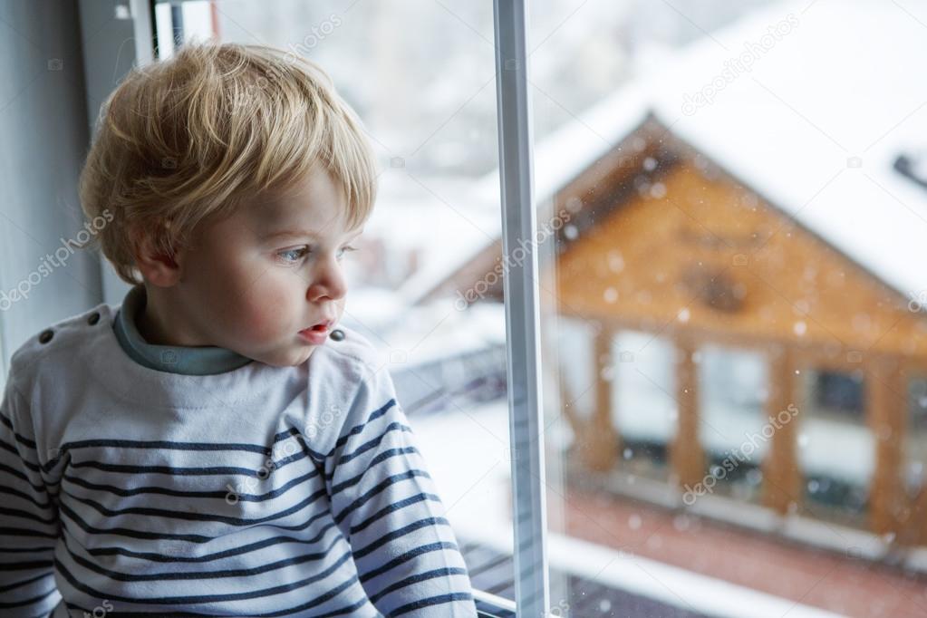 Little toddler boy looking out of the window on winter day with