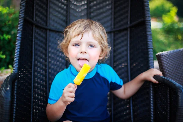 Bonito menino loiro comendo sorvete amarelo, ao ar livre — Fotografia de Stock