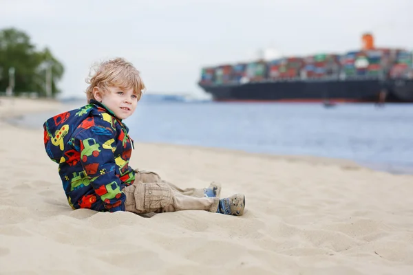 Pequeño niño sentado en la playa de arena y mirando en containe — Foto de Stock