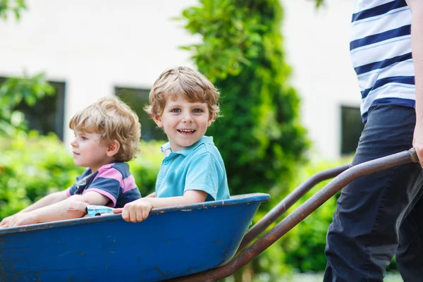 Two little boys having fun in wheelbarrow pushing by father — Stock Photo, Image