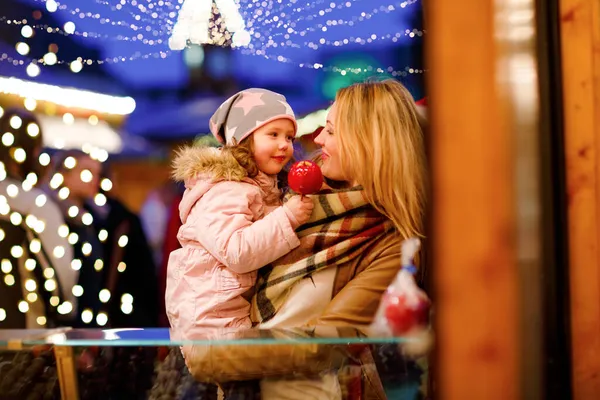 Cute little preschool girl and young mother eating crystallized sugared apple on German Christmas market. Happy family, girl and woman with lights on background. Mum holding daughter on arms. — Stock Photo, Image