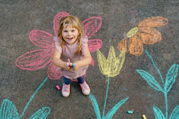 Liten förskola flicka målning med färgglada kritor blommor på marken på bakgården. Positivt glad barn rita och skapa bilder på asfalt. Kreativa utomhus barn aktivitet på sommaren. — Stockfoto