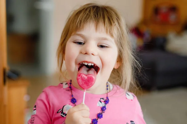 かわいい幼児の女の子とお菓子。愛らしい子供食べるとピンク色のハートロリポップをなめる、屋内. — ストック写真