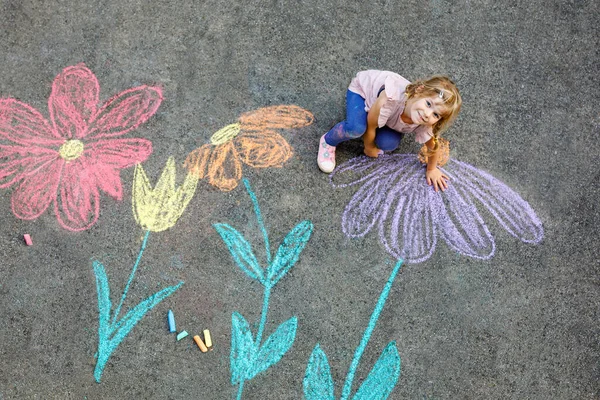 Μικρή προνήπιο κορίτσι ζωγραφική με πολύχρωμα κιμωλίες λουλούδια στο έδαφος στην πίσω αυλή. Θετικό, χαρούμενο παιδί ζωγραφίζει και δημιουργεί εικόνες στην άσφαλτο. Δημιουργική υπαίθρια παιδική δραστηριότητα το καλοκαίρι. — Φωτογραφία Αρχείου
