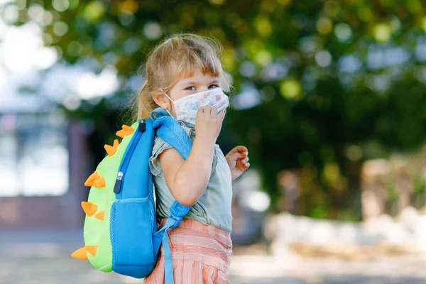 Kleines Kleinkind an ihrem ersten Schultag mit medizinischer Maske gegen Coronavirus. Gesunde schöne Baby zu Fuß in die Vorschule und den Kindergarten. Glückliches Kind mit Rucksack — Stockfoto