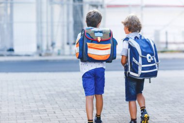 Sırt çantası ya da çantası olan iki küçük çocuk. Okulun yolu üzerindeki okul çocukları. Sağlıklı sevimli çocuklar, erkek kardeşler ve en iyi arkadaşlar sokakta evden ayrılıyor. Okula dönelim. Mutlu kardeşler..