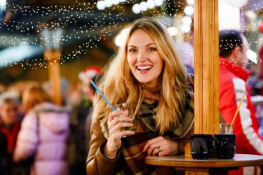 Sıcak punç içen, Alman Noel pazarında sıcak şarap içen genç bir kadın. Arka planda arkadaşları ve ışıkları olan kışlık elbiseli mutlu bir insan. Aile, gelenek, tatil konsepti.