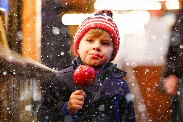 Cute little smiling preschool boy on German Christmas market. Happy child in winter clothes eating sweet sugared glazed xmas apple on with lights on background. Family, tradition, celebration concept — Stock Photo, Image