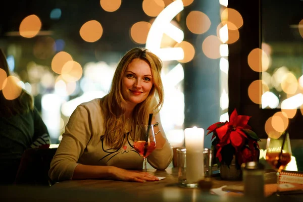 Young woman drinking Aperol Spritz cocktail after work in an indoor pub bar and restaurant. Christmas market with ferris wheel and lights on background. Happy woman dreaming on evening or night