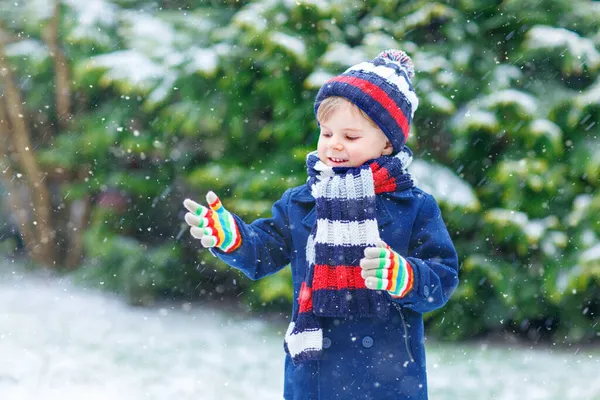 Aranyos kis vicces gyermek színes téli divat ruhák szórakozás és játszik a hó, szabadban hóesés alatt. Aktív szabadtéri kikapcsolódás gyerekekkel. Gyerek fiú és kisgyerek hópelyheket fog. — Stock Fotó