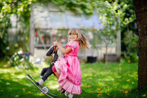 Petite fille préscolaire étreignant avec jouet de cheval à bascule. Joyeux enfant en robe de princesse le jour ensoleillé d'été dans le jardin. Fille en amour avec son favori vieux jouet vintage animal. — Photo