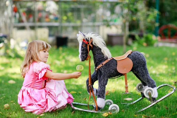 Dziewczynka z przedszkola przytulająca się do kołyszącej zabawki. Szczęśliwe dziecko w sukience księżniczki na słoneczny letni dzień w ogrodzie. Dziewczyna zakochana w swoim ulubionym starym zwierzęciu vintage zabawki. — Zdjęcie stockowe
