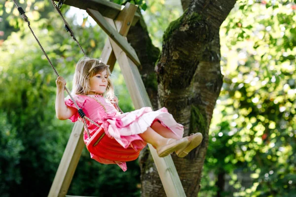 Menina pequena feliz que se diverte no balanço no jardim doméstico. Criança sorridente em vestido de princesa balançando no dia ensolarado. Menina pré-escolar rindo e chorando. Lazer ativo e atividade ao ar livre. — Fotografia de Stock