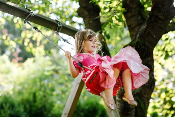 Szczęśliwa dziewczynka bawiąca się na huśtawce w ogrodzie. Uśmiechnięte dziecko w sukience księżniczki huśtające się w słoneczny dzień. Dziewczyna z przedszkola śmieje się i płacze. Aktywny wypoczynek i aktywność na świeżym powietrzu. — Zdjęcie stockowe
