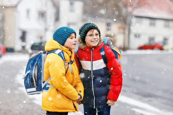 Två små pojkar i grundskolan som går till skolan under snöfallet. Glada barn som har roligt och leker med första snön. Syskon och bästa vän med ryggsäck i färgglada vinterkläder. — Stockfoto